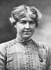 Monochrome photograph portrait of a woman in her twenties, shown from the bust upward, the woman wearing a vertical-patterned blouse decorated by a row of buttons between the shoulders and the closed collar, her face directly forward gazing at the viewer, her cheeks prominent and fleshy, the mouth slightly opened in a tight smile, the coarse, sun-bleached sandy-coloured hair parted in the middle, extending to the ears in an overall loose wave with flyaway strands