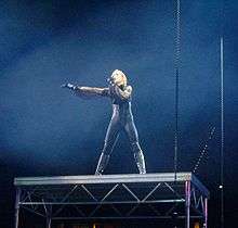 A blond woman stands on a steel tabletop. She wears a tight, body-hugging dress, accompanied by boots and gloves. She sings to a microphone in her left hand and her right hand is stretched out. The table is held by steel cables.