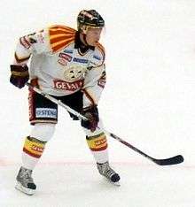 An ice hockey player leaning forward with his ice hockey stick resting on the ice. He is wearing a black helmet and a white and black uniform.