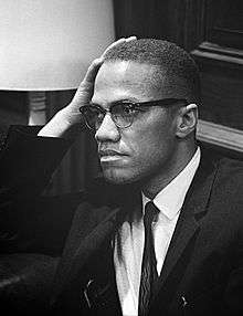 An African-American man in his forties, wearing glasses and a suit and tie, sitting and looking to the right, with his hand resting on his right temple.