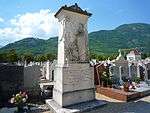 A grave marker about three meters tall with a classical motif and wooded hills in the background