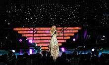 A blonde woman wearing a white gown sings. Behind her are brightly lit stairs, and twinkling lights are on the ceiling.