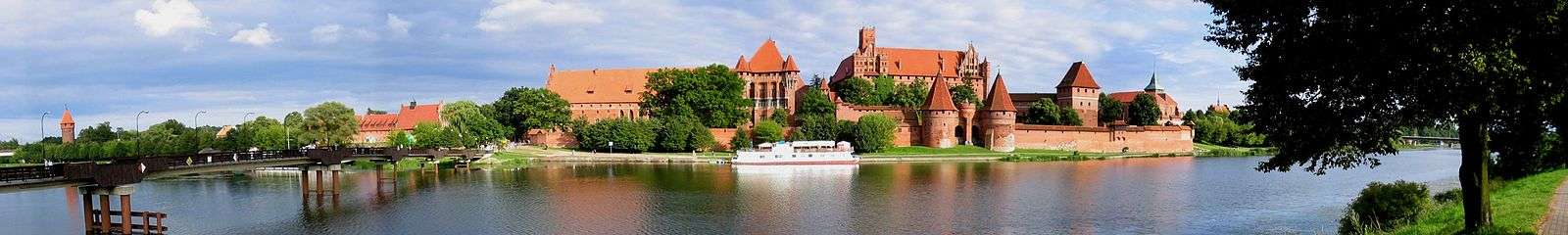 An orange brick castle with a curtain wall and a central keep. The site is surrounded by water. The gateway is flanked by two round towers with high peaked roofs. Aside from the keep, there is another building within the castle rising above the curtain wall.