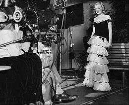 Monroe wearing a dress and facing a movie camera in a publicity photograph taken during her first film contract.