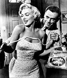 Monroe in The Seven Year Itch. She is holding a bag of chips and wearing a dress, which shoulder straps are undone. Behind her is Tom Ewell, who is holding the straps.