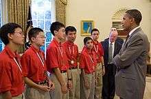 Six people in red shirts and beige pants standing in a line next to men in black suits all in front of a man wearing a grey suit and a red tie