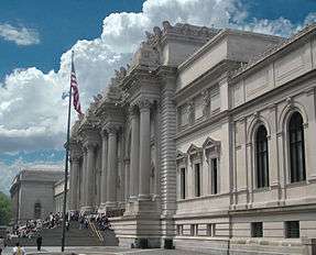 Facade of imposing building with Greek columns. Large colored banners hang from the building's top. A crowd of people is in front.