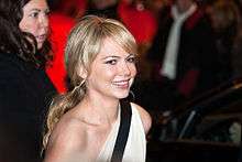 An image of a smiling Caucasian woman. She has her blonde hair in a ponytail and is wearing a one shouldered white dress with a black horizontal stripe.