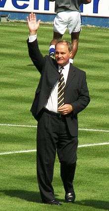 A man in a suit with his right hand raised, standing on a football pitch.