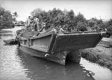 Four shirtless men in a large barge moored at a river bank. The barge has a ramp at the front of a catamaran-like hull.