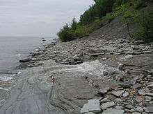 A stream spills over layered rock on a lake shore covered with small flat rocks