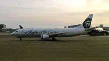 Left side view of an airplane taxiing on the tarmac. Buildings and other aircraft visible in background.