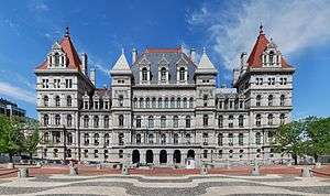 An ornate building, several stories high, of light colored stone. Many arches are visible on its front. On its sides are two large towers with pyramidal red roofs, echoed by similar smaller towers closer to the center with stone tops. In front of the camera, at bottom, is a plaza with a wavy-line pattern.