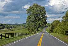 A two-lane road stretching out ahead of the camera to a distant curve to the right. On the left is a rolling of fields, woods and two small houses with a brown fence running alongside the road