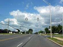 A four-lane highway proceeds into the background, where it disappears from view after descending a hill. Buildings home to various businesses, streetlights, and sidewalks line both sides of the highway. Two overhead signs held up by wires instruct drivers destined for NY 22B southbound to continue straight and those destined for NY 3 westbound to turn right ahead.