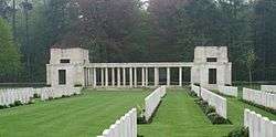 A grass lawn divided by rows of white grave stones. A pair of block-like white stone buildings flank a range of columns.