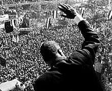 The back of a man waving to the throng below