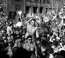 A man standing in an open-top vehicle and waving to a crowd of people surrounding the vehicle. There are several men seated in the vehicle and in another trailing vehicle, all dressed in military uniform