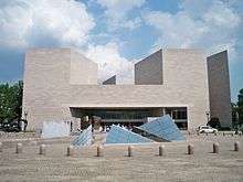 A large grey building rises above a stone plaza. Short square towers appear on either side of the building, and an array of irregular glass pyramids are in the middle of the plaza.