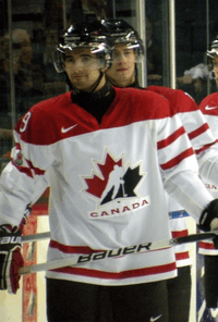 Hockey player in red and white Canada uniform. He has a half-smile on his face and holds his stick in his hands.