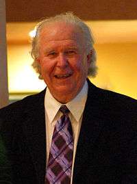 A man with disheveled light gray hair wearing a black suit jacket and purple striped tie smiles and looks directly ahead.