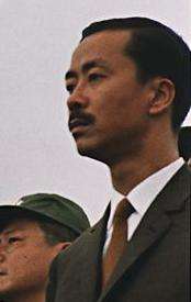 Middle-aged man with side-parted  black hair and moustache, in a black suit, white shirt and brown tie. To the left is a clean-shaven Asian man with black hair and a green military cap.