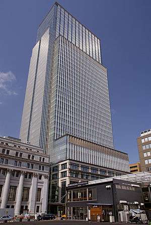 Ground-level view of a rectangular, glass high-rise; adjoining the high-rise is a stone building featuring columns