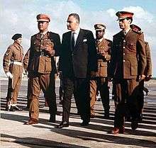 Three men walking side-by-side. The man in the middle is wearing a suit, while the two to his side are wearing military uniforms and hats. There are a few other men in uniform walking behind them