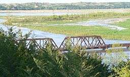 River flowing through marshy lowlands; seen from high bank, past conifers