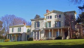 An elaborate yellow house with porches, a square tower on the right and green wooden shutters seen from slightly below it.