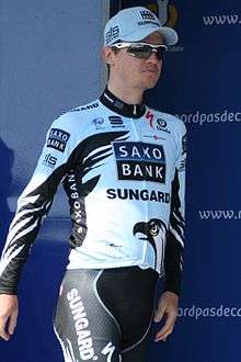 A man of about thirty wearing a black and pale blue cycling jersey with white trim, shades, and a white baseball cap walks by a blue backdrop.