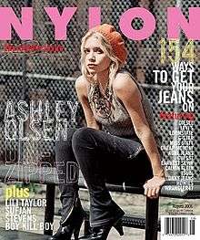 Nylon magazine cover, with the name in all caps block bold Helvetica font, featuring Ashley Olsen seated on a walkway railing in black skinny jeans with boot flares, large-bead necklace, a grey tank top, and orange knit cap, in front of a chain link fence.