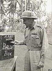 Three-quarter-length outdoor portrait of moustacioed man in light tropical military uniform with pith helmet and pilot's wings on left breast pocket, placing folded paper into a box marked "RAAF BALLOT BOX"