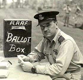 Man in military uniform with peaked cap seated at a desk beside a box marked "RAAF Ballot Box"