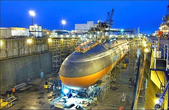 Black submarine with orange paint from cheatline down in drydock at nightfall.