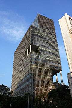 Ground-level view of a 20-storey skyscraper with a brown facade; the building has a rectangular cross section and is cut-off roughly 10 stories up, with the uppermost 10 floors partially protruding out over the street below.