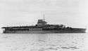 Side view of a gray anchored aircraft carrier with a short flight deck. Her island incorporates a funnel and is surmounted by a tall mast.