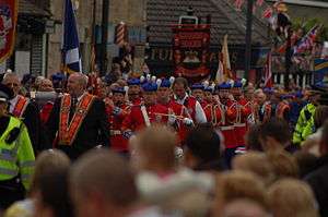 A street is occupied by men and women in formal uniform, some bearing flags, some bearing banners, some playing musical instruments.