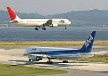 A white and red-tailed Japan Airlines aircraft above runway, with landing gears down, and an All Nippon Airways in blue and white livery taxiing.