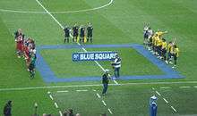 Two football teams line up opposite each other on the halfway line, with the referee, assistants and the fourth official standing to their side