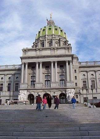 Looking up from a large, stone staircase is a marble facade of a building with a large, pale green dome. Several people are walking down the stairs.