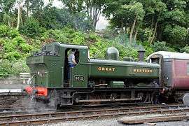 A pannier tank locomotive is standing at a platform with a red passenger carriage in front of it. The locomotive is mainly painted green above the running plate, although the chimney is black and the safety valve cover is polished brass. The lettering "GREAT WESTERN" is shown in yellow with red shading on the side of the pannier tank. The rear buffer beam is painted red.