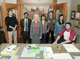 A group of people stand in an office-style room in front of a table with documents and booklets. From left to right stand a man with arms crossed wearing a black shirt, a man wearing a gray suit and green tie with his arms behind his back, a woman in a gray suitcoat and red shirt, a woman with a gray jacket and purple shirt, a man with arms crossed wearing a tan suit, a young girl wearing a blue blouise and gray shirt and a seated man wearing a white T-shirt with red sleeves.