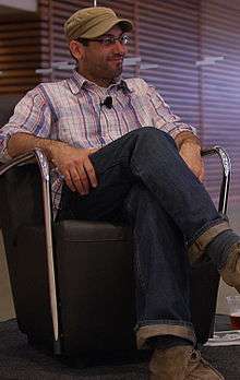 A man with hat and glasses sitting on a chair and crossing his legs.