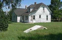 One-and-a-half story wood house with peeling paint; in foreground, door leading to storm cave