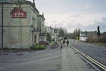 Gray stone building on the left with a pub sign outside it. A road is central to the picture with a white coloured building on the right.