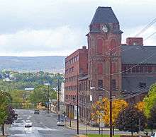 A brick tower with peaked roof on the right-hand side of a street in front of the camera. There are green hills in the background.