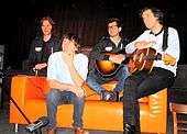 Four men sitting on an orange sofa, two holding guitars and one behind keyboards that are set on the left arm of the sofa.