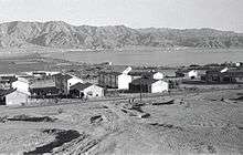 A black and while photograph of Eilat taken from the West. The settlement is small with only a few buildings. In the distance, the Jordanian settlement of Aqaba is visible, and backdropped by mountains.