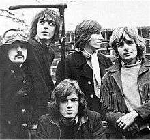 A black and white photo of five men standing in front of a brick wall.